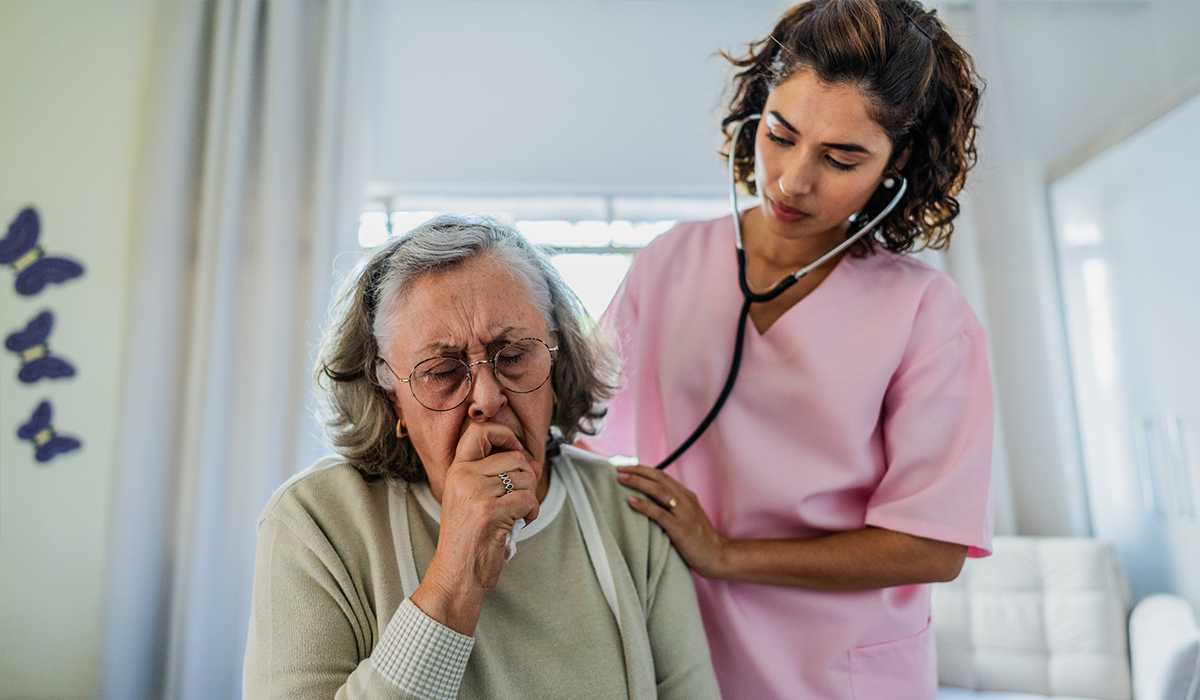 Image of an elderly woman coughing and a healthcare worker pressing a stethoscope to her back